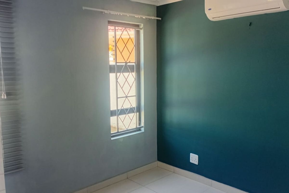 An empty bedroom featuring tiled flooring, a wall-mounted air conditioning unit, and a window fitted with security bars and blinds.