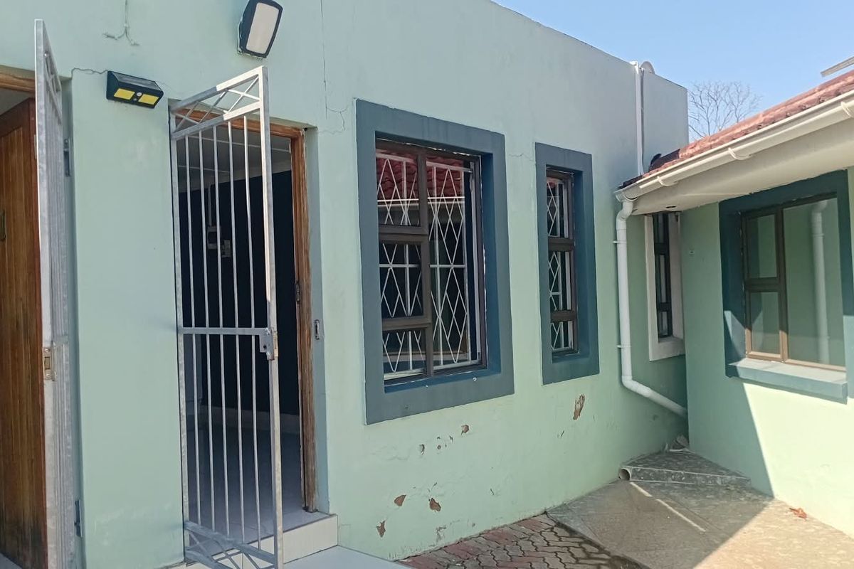 Rear exterior view of the property featuring a security gate, window security bars, and paved ground area with visible exterior wall paint deterioration.