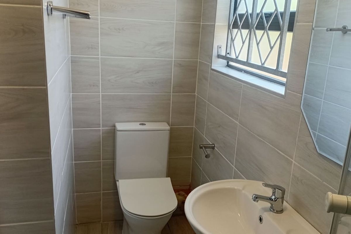 A clean, modern bathroom featuring neutral wood-look wall tiles, a white toilet, a pedestal washbasin, and a window with security bars.