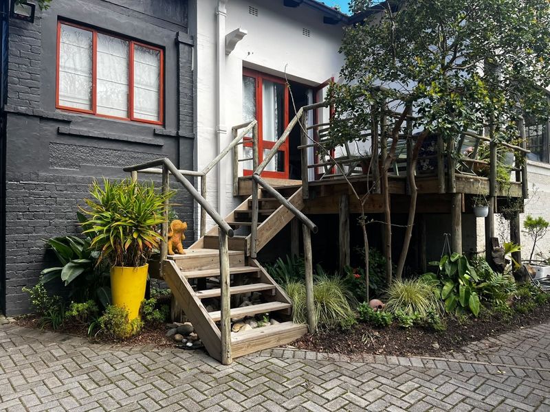 Rear exterior view of the cottage featuring a raised timber deck with staircase access, paved ground area, and painted brick facade.