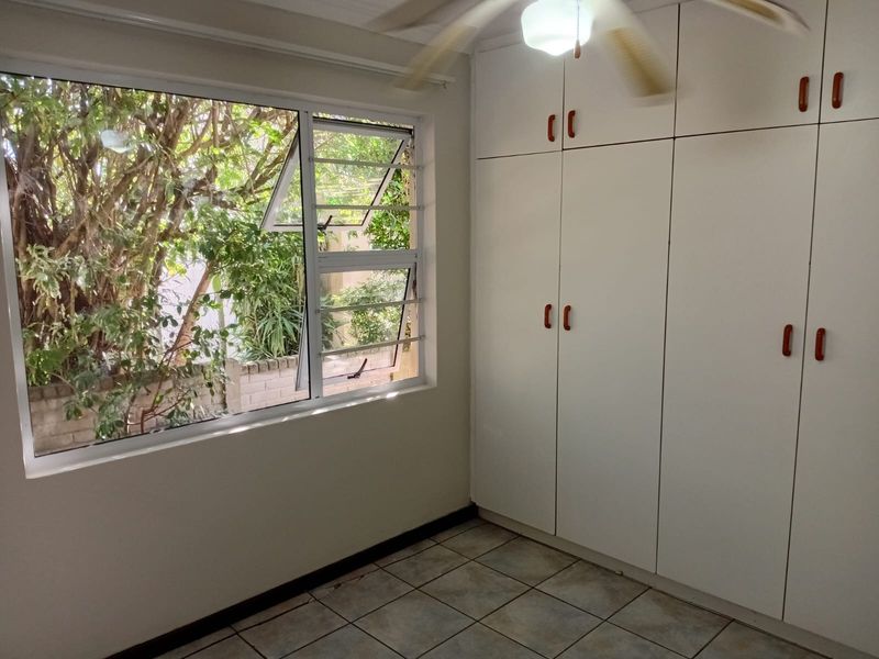 A bright, empty bedroom featuring tiled flooring, built-in cupboards, and a ceiling fan, with large windows overlooking the garden.
