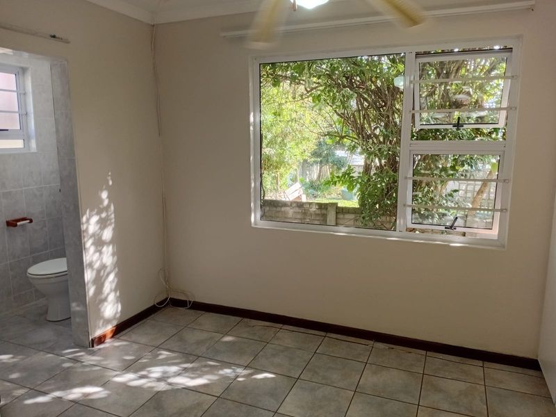 An empty bedroom featuring tiled flooring, a ceiling fan, and an en-suite bathroom, with large windows providing natural light and garden views.