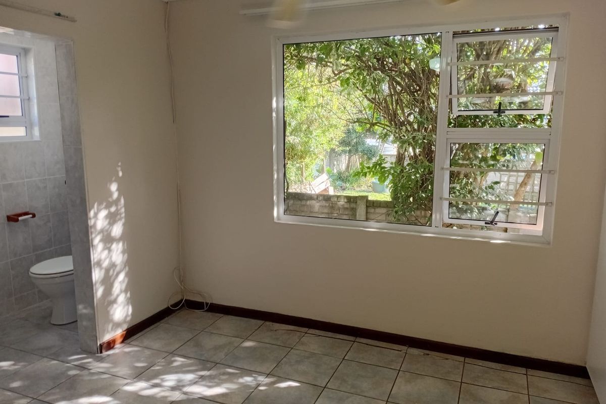 An empty bedroom featuring tiled flooring, a ceiling fan, and an en-suite bathroom, with large windows providing natural light and garden views.