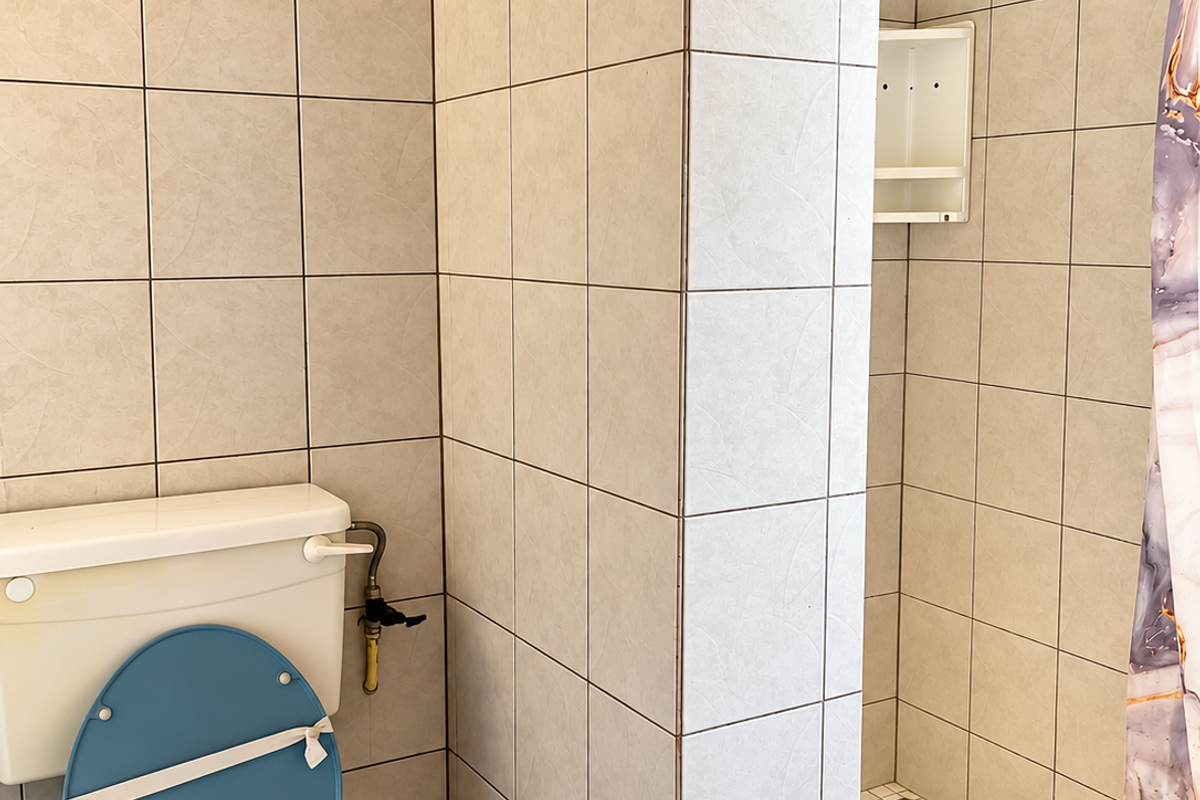 A functional bathroom featuring a toilet and a tiled shower enclosure with built-in shelving, requiring cosmetic updates to modernize the space.