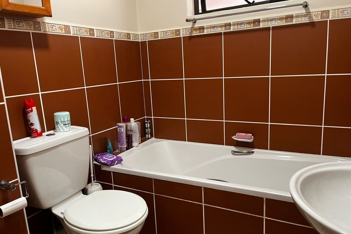 A functional bathroom featuring a built-in bathtub, pedestal basin, and toilet, finished with dark brown ceramic wall tiles and a window with security bars.