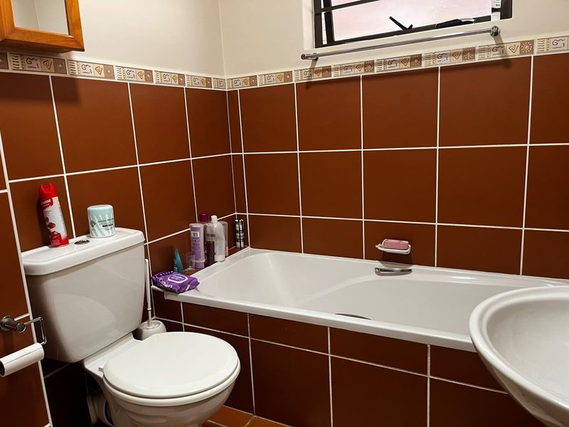 A functional bathroom featuring a built-in bathtub, pedestal basin, and toilet, finished with dark brown ceramic wall tiles and a window with security bars.