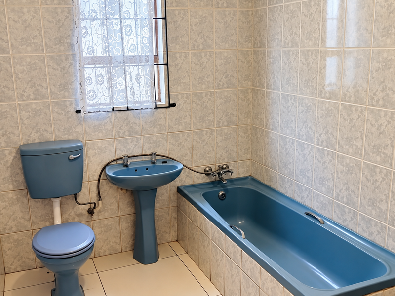 A dated bathroom featuring blue sanitaryware, including a pedestal basin, toilet, and bathtub, complemented by full-height wall tiling and a window with security bars.