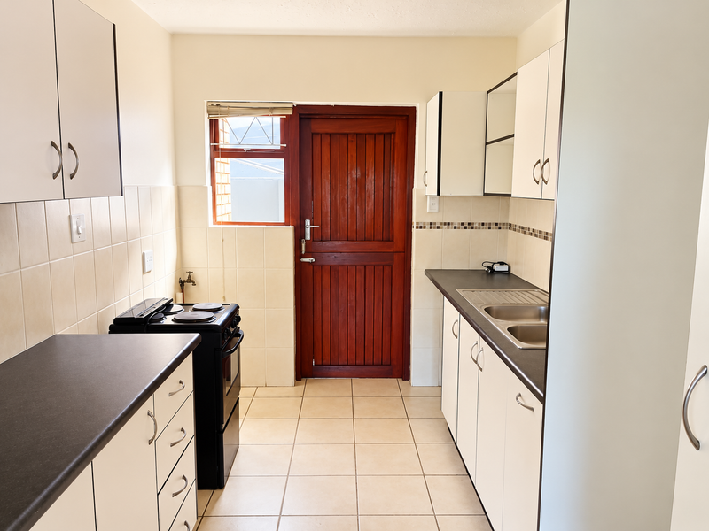 A functional galley-style kitchen featuring white cabinetry, dark laminate countertops, a double stainless steel sink, and a freestanding stove with direct exterior access.