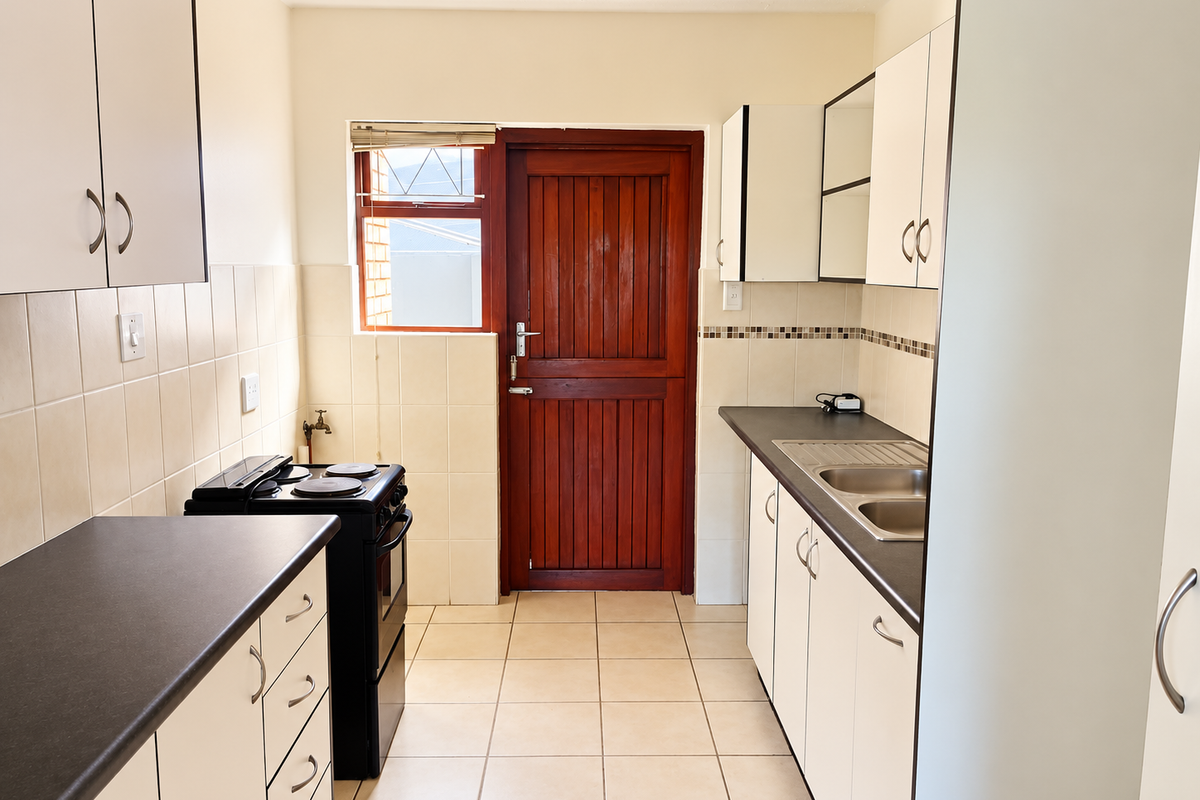 A functional galley-style kitchen featuring white cabinetry, dark laminate countertops, a double stainless steel sink, and a freestanding stove with direct exterior access.