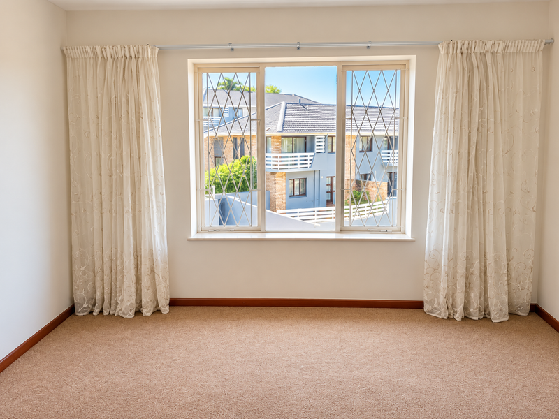 A bright, empty bedroom featuring neutral carpeting, clean walls, and a large window fitted with security bars and curtain rods.