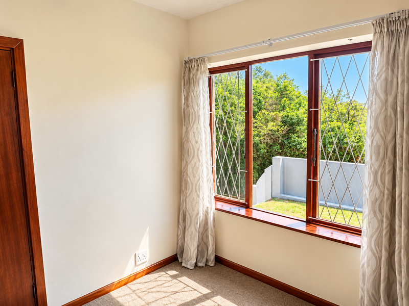 A bright, empty bedroom featuring wooden window frames with security bars, carpeted flooring, and neutral walls, offering a pleasant view of the garden.