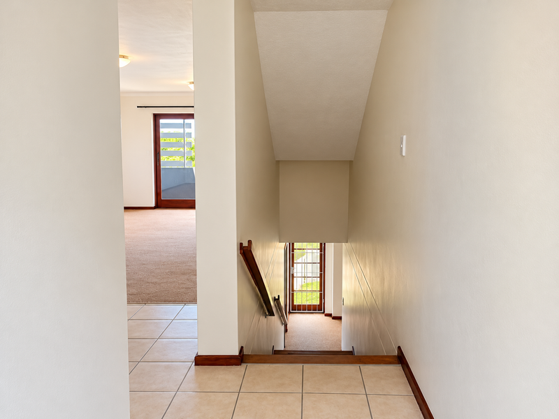 A bright hallway featuring a staircase with a wooden handrail, connecting tiled and carpeted living areas with access to an exterior door.