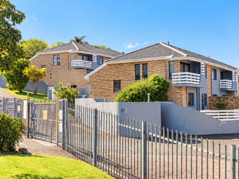Exterior view of a face brick townhouse complex featuring multiple balconies, tiled roofing, and secure metal perimeter fencing with a paved driveway.