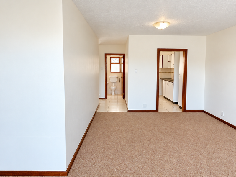 An empty living area featuring neutral carpeting, leading into a functional kitchen and a separate bathroom with tiled flooring.