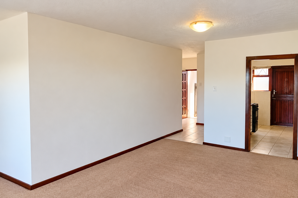 A spacious, empty living area featuring neutral carpeting, clean white walls, and tiled flooring leading into an adjacent room with wooden door frames.