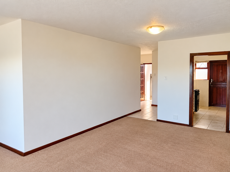 A spacious, empty living area featuring neutral carpeting, clean white walls, and tiled flooring leading into an adjacent room with wooden door frames.