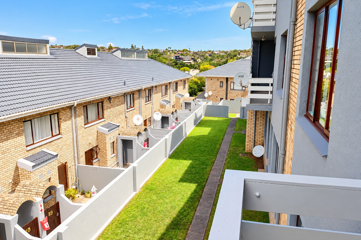 An elevated view of the rear exterior of a townhouse complex, featuring brick facades, tiled roofs, and a shared green space with a paved walkway.