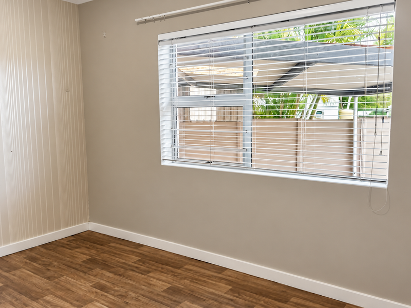 A bright, empty bedroom featuring modern laminate flooring, neutral wall tones, and fitted venetian blinds, offering a clean canvas for personalization.
