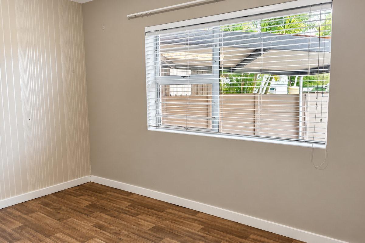 A bright, empty bedroom featuring modern laminate flooring, neutral wall tones, and fitted venetian blinds, offering a clean canvas for personalization.