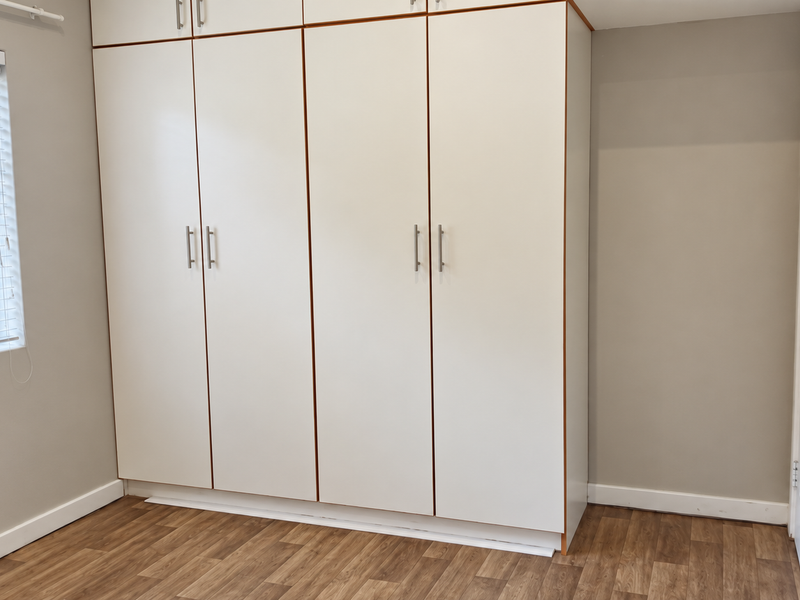 A clean, empty bedroom featuring modern built-in cupboards, laminate flooring, and a window with venetian blinds.