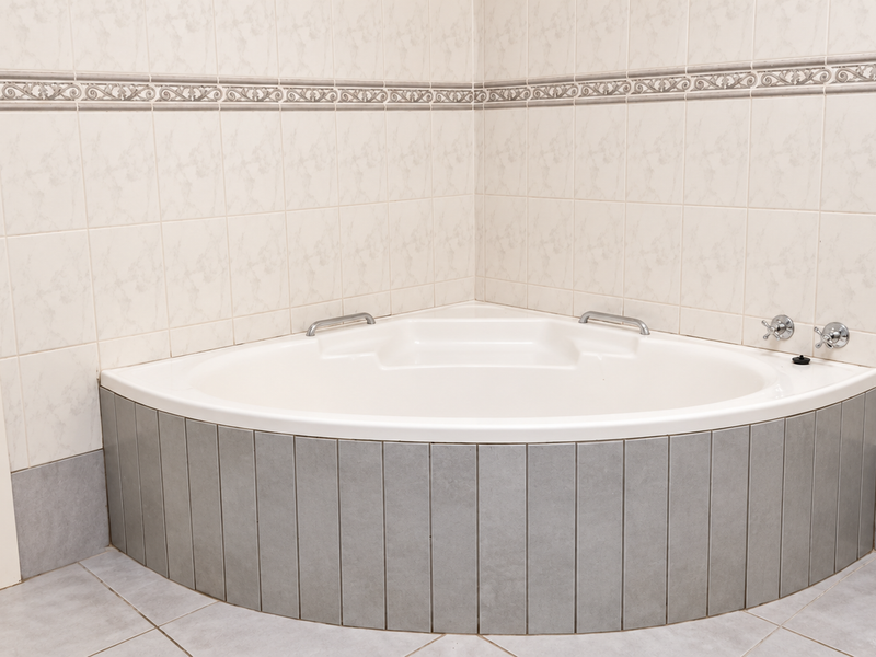 A clean bathroom featuring a corner bathtub with chrome fixtures and neutral wall and floor tiling.