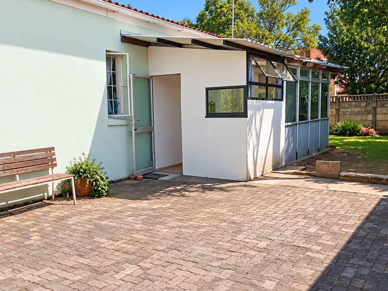 Rear exterior view featuring a paved patio area, a small enclosed sunroom extension, and a concrete boundary wall in a garden setting.