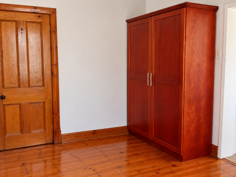 A bedroom featuring polished timber flooring, matching timber door and architraves, and a built-in wardrobe, with an adjacent view into a tiled bathroom.