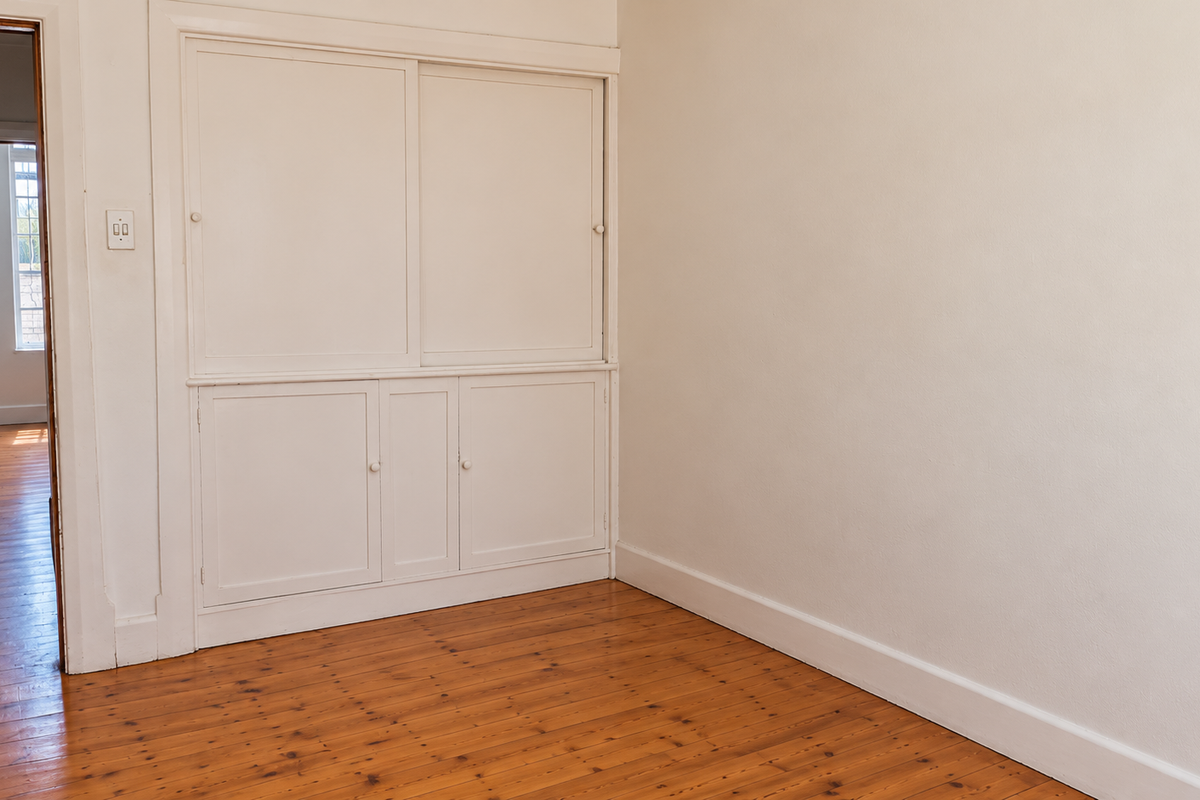 A bright, empty bedroom featuring polished timber flooring and classic built-in storage cabinetry, with a view into an adjacent room.
