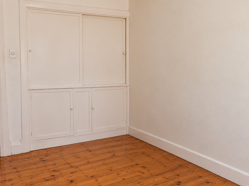 A bright, empty bedroom featuring polished timber flooring and classic built-in storage cabinetry, with a view into an adjacent room.
