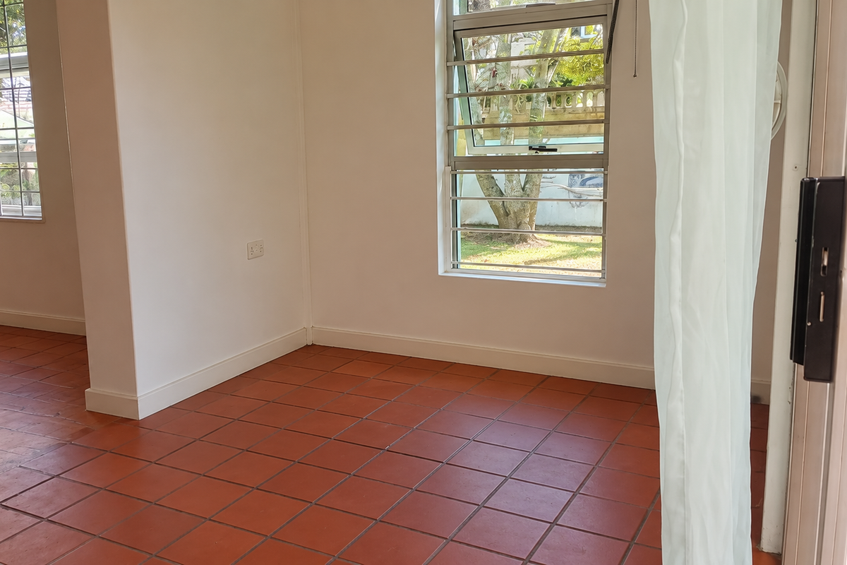 An empty living area featuring terracotta floor tiles, white walls, and aluminium windows with security bars, offering a bright and neutral blank canvas.