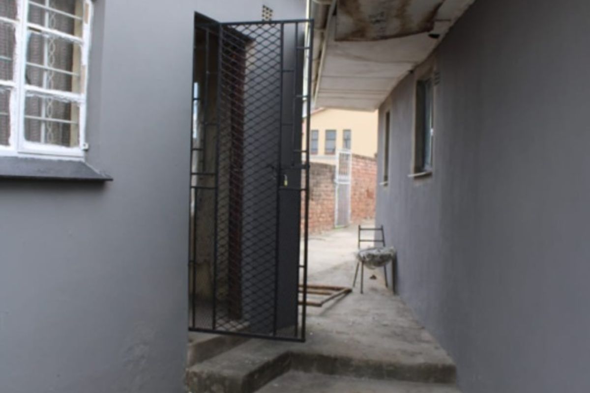 A narrow exterior side passage featuring concrete steps, security gates, and painted masonry walls with visible signs of weathering and maintenance requirements.