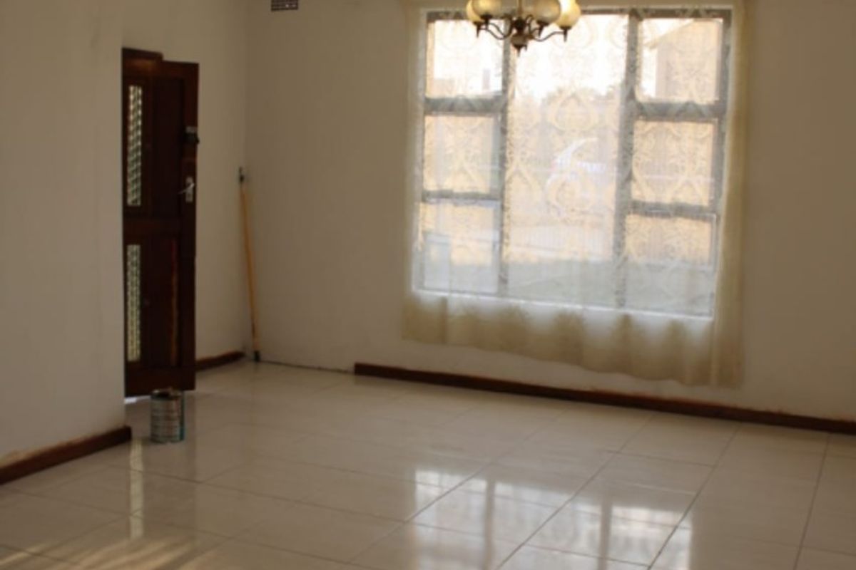 A spacious, empty living room featuring neutral tiled flooring, a large window with natural light, and a classic chandelier fixture.