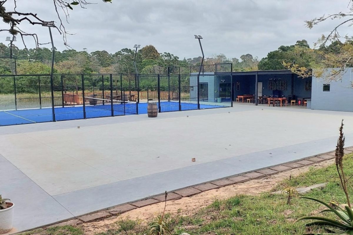 Outdoor sports facility featuring a padel court with floodlights, adjacent to a paved patio and covered entertainment area.