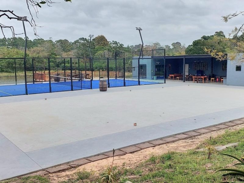 Outdoor sports facility featuring a padel court with floodlights, adjacent to a paved patio and covered entertainment area.
