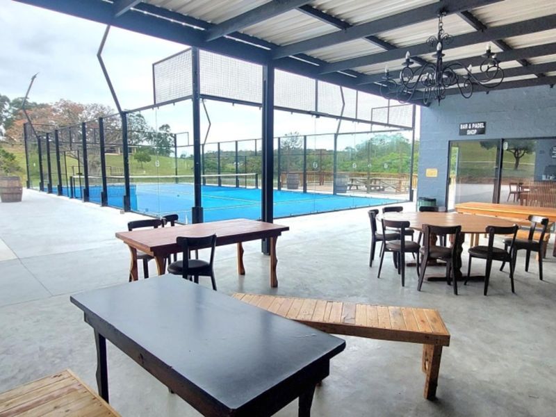 A covered outdoor patio area overlooking a professional padel court, featuring concrete flooring, a metal roof structure, and integrated lighting.