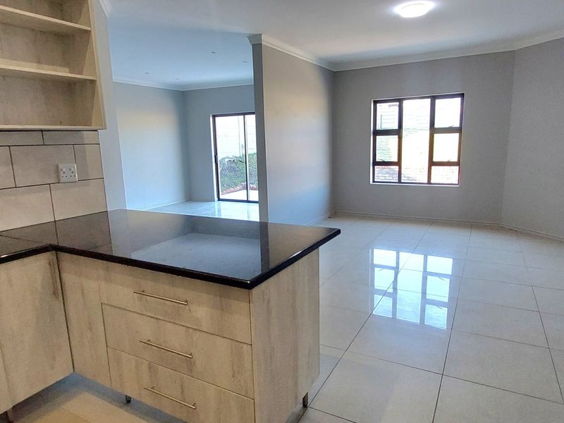 An open-plan living and kitchen area featuring light wood-finish cabinetry, granite countertops, tiled flooring, and a sliding door leading to the exterior.