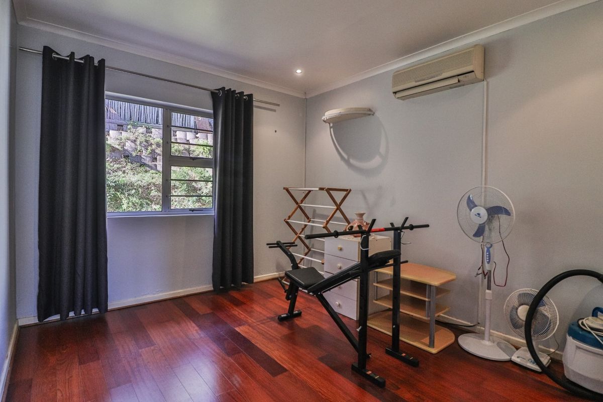 A bedroom featuring polished timber flooring, a wall-mounted air conditioning unit, and a window with views of the exterior retaining wall.
