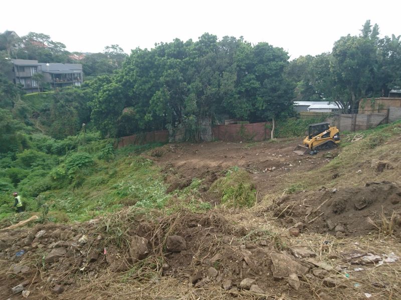 A sloped residential plot currently undergoing site clearing and earthworks, featuring mature trees and surrounding vegetation.