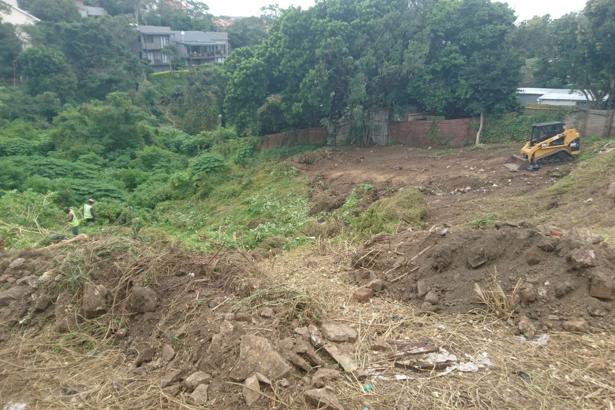 A sloped residential plot currently undergoing site clearing and earthworks, featuring dense surrounding vegetation and a retaining wall structure.