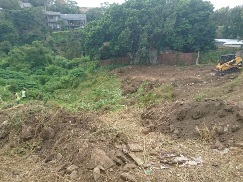 A sloped residential plot currently undergoing site clearing and earthworks, featuring dense surrounding vegetation and a retaining wall structure.