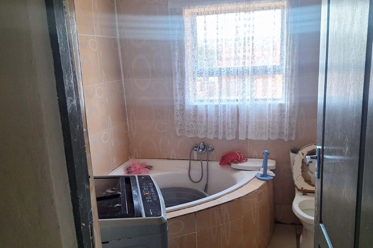 A compact bathroom featuring a corner bathtub, toilet, and tiled walls, with space currently utilized for laundry equipment.
