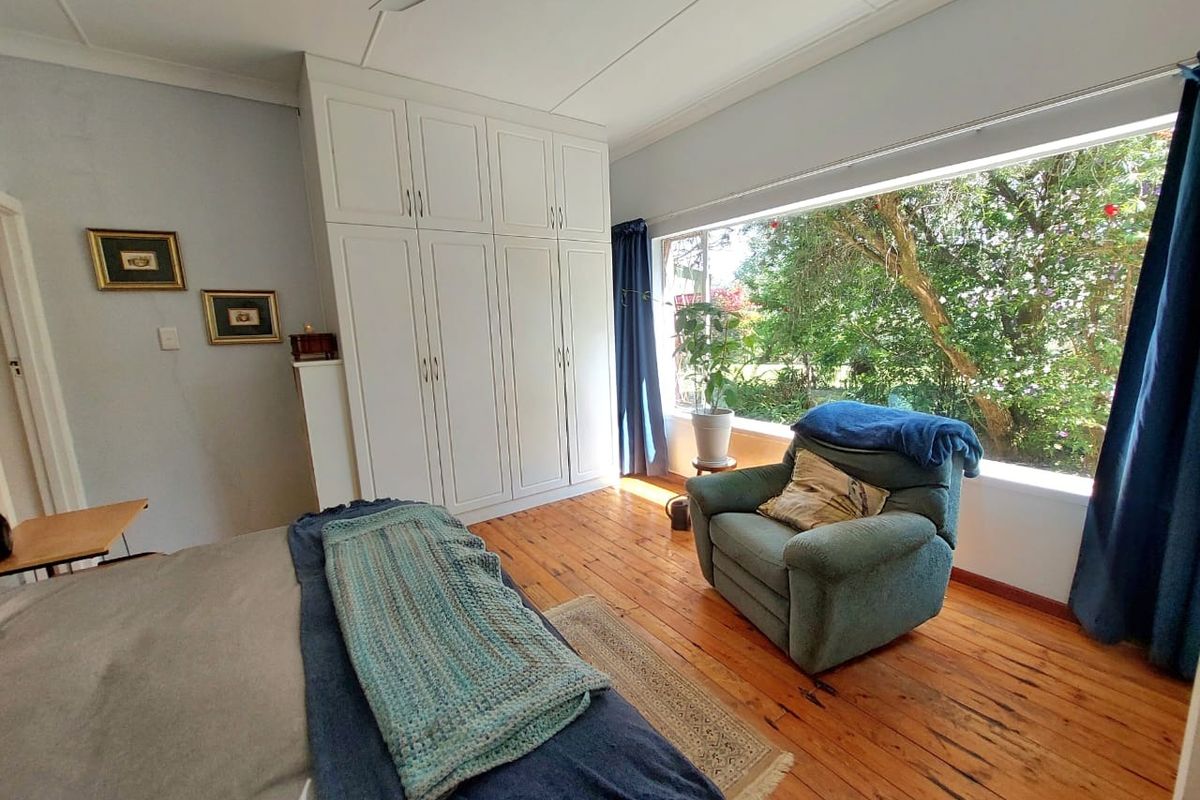 A bedroom featuring polished timber flooring, built-in cabinetry, and a large window providing natural light and garden views.
