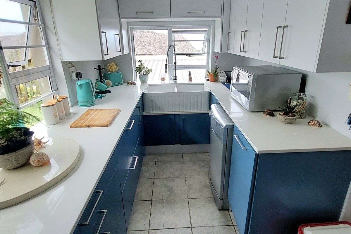 A compact, modern kitchen featuring white upper cabinetry, blue lower cabinets, white countertops, and a farmhouse-style sink with a window view.