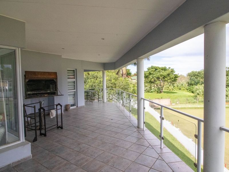 Spacious covered balcony featuring tiled flooring, a built-in braai, and a glass balustrade overlooking a green landscape.