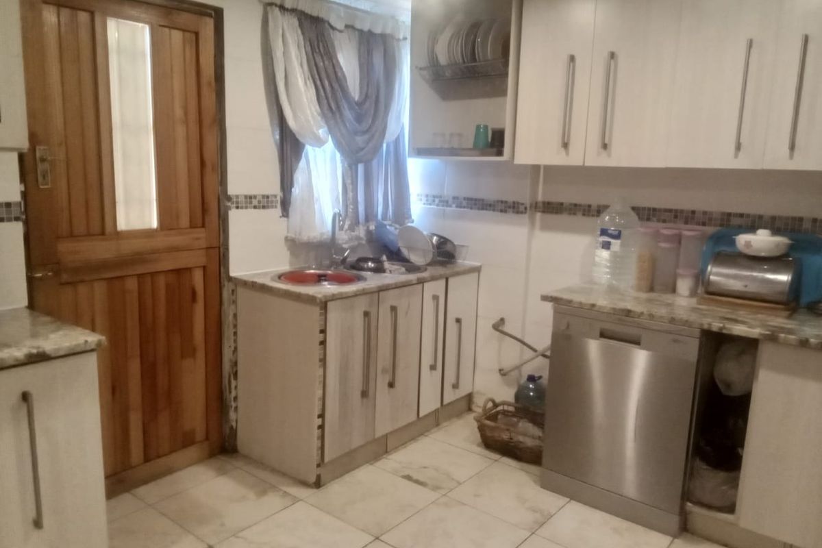 A functional kitchen featuring built-in cabinetry, tiled flooring, a stainless steel sink, and an integrated dishwasher, with a wooden door leading to the exterior.