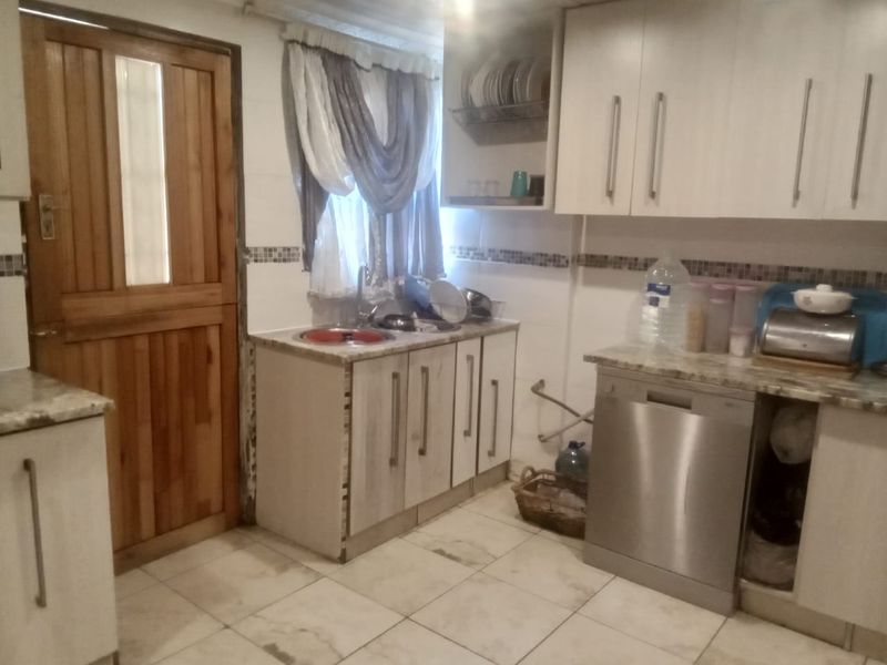 A functional kitchen featuring built-in cabinetry, tiled flooring, a stainless steel sink, and an integrated dishwasher, with a wooden door leading to the exterior.