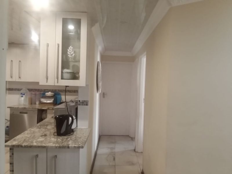 A functional kitchen area featuring granite countertops, white cabinetry with glass-front detailing, and a mosaic tile backsplash, leading into a hallway.