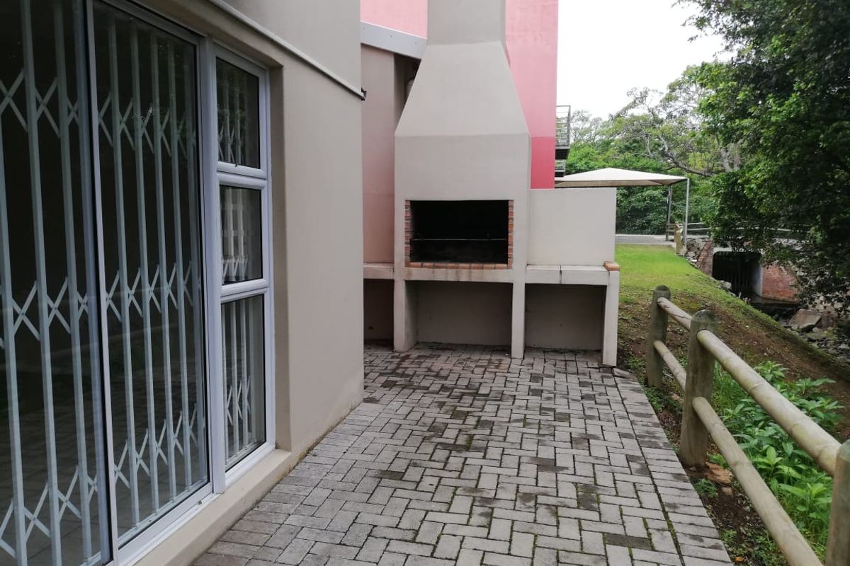 A paved outdoor patio area featuring a built-in brick braai, accessible via a sliding door with a security gate, overlooking a garden space.