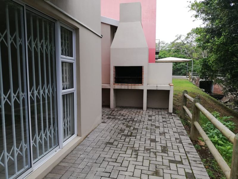 A paved outdoor patio area featuring a built-in brick braai, accessible via a sliding door with a security gate, overlooking a garden space.