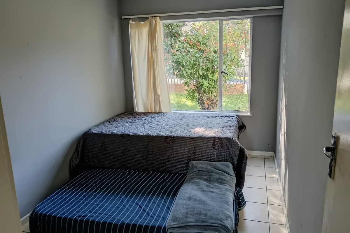 A compact bedroom featuring tiled flooring and a large window with security bars, offering natural light and a view of the garden.